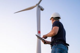 Male engineer in the helmet controls and maintains the operation of a wind turbine using a tablet on the background of windmill.