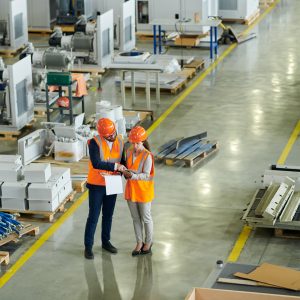 High angle full length portrait of bearded businessman wearing hardhat inspecting production workshop accompanied by female factory worker, copy space